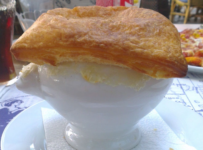 Close-up of a pastry-topped soup bowl on a saucer with a pizza slice in background