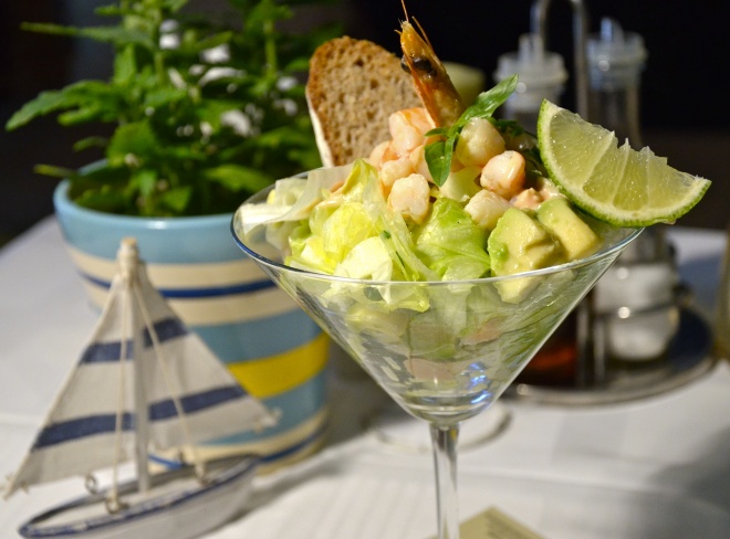 Martini glass filled with shrimp and avocado salad garnished with lime, bread, and shrimp, with decorative sailboat
